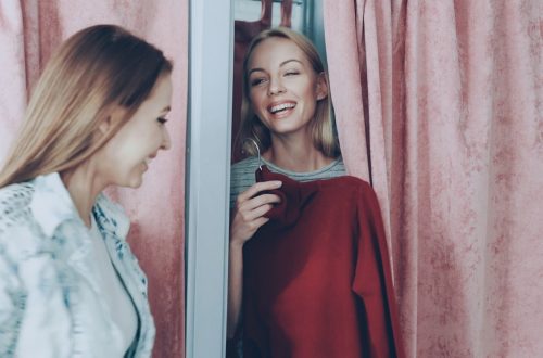 Young blond woman half hidden behind curtain in fitting room in a store asking to fetch her red knitted dress. Female friend bringing her clothes, and she drawing curtain to try it on