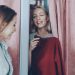 Young blond woman half hidden behind curtain in fitting room in a store asking to fetch her red knitted dress. Female friend bringing her clothes, and she drawing curtain to try it on